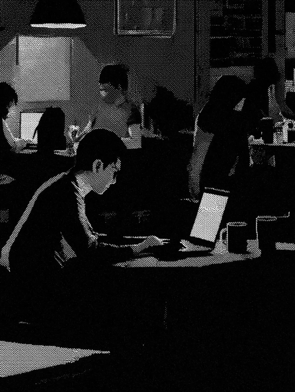 A dim room at night, several people working at laptops under a low hanging lamp, screens the brightest thing in the picture.